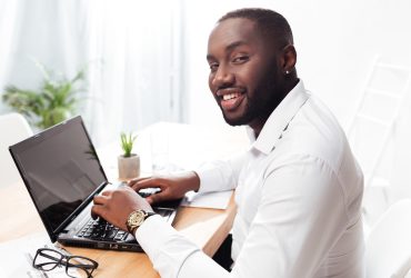 Portrait of smiling african american businessman in white shirt sitting and happily looking in camera while working on his laptop in office isolated