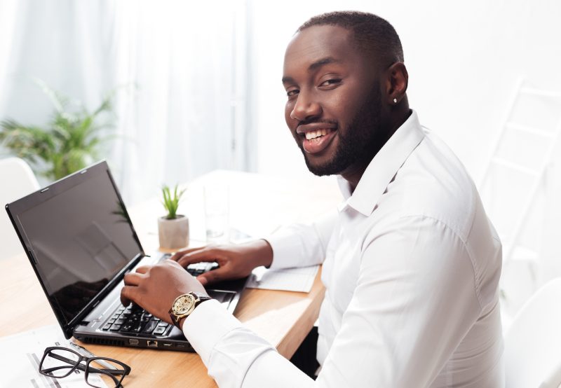 Portrait of smiling african american businessman in white shirt sitting and happily looking in camera while working on his laptop in office isolated